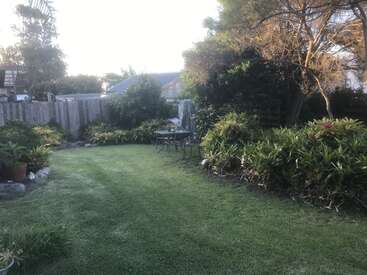 The image depicts a serene backyard with a lush green lawn, surrounded by trees and bushes, featuring a patio area with a table and chairs in the center.