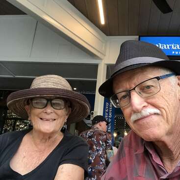 Das Bild zeigt ein älteres Paar, das zusammen ein Selfie macht, beide mit Hut und Brille, mit einem Schild im Hintergrund und einem Mann in einem Hawaiihemd hinter ihnen.