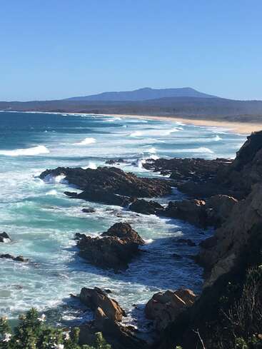 The image depicts a rugged coastline with dark rocks, white waves, and a sandy beach, set against a backdrop of a mountain range under a clear blue sky.