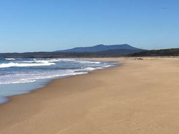 The image depicts a serene beach scene with gentle waves, a sandy shore, and a distant mountain range under a clear blue sky on a sunny day.