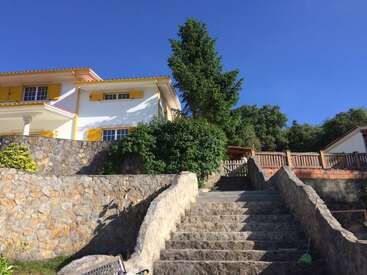 L'image représente une maison blanche à deux étages avec des accents jaunes, avec un mur de pierre et un escalier menant à l'entrée, sur fond de ciel bleu clair.
