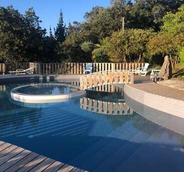 Une piscine paisible, agrémentée d'un petit îlot circulaire, d'une passerelle en bois et de plusieurs chaises longues, est entourée d'arbres, de verdure et d'une clôture en bois.