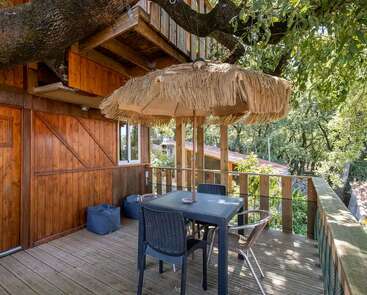 Une terrasse en bois chaleureuse, entourée d'arbres luxuriants, équipée d'une table carrée avec quatre chaises, d'un parasol en paille et d'un coin salon extérieur propice à la détente à l'ombre.