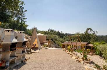 Cet espace extérieur comprend trois barbecues en pierre, une balançoire en bois, une tente tipi, une cabane, un sol recouvert de gravier et une végétation luxuriante sous un ciel dégagé.