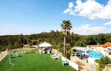 Un magnifique espace extérieur comprenant du gazon synthétique, des chaises longues, un kiosque, un palmier, un jardin clôturé, des chaises bleues, une piscine à débordement et une pergola, le tout entouré d'une vue sur une forêt verdoyante.