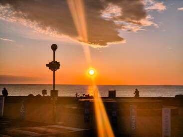 Um belo pôr do sol sobre o oceano, com tons alaranjados vibrantes, nuvens dramáticas, silhuetas de pessoas, um poste de luz e um raio de sol dourado atravessando a imagem.