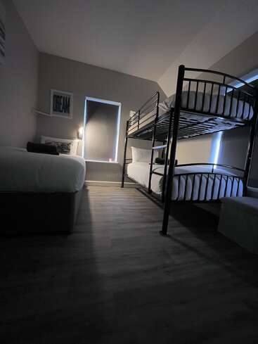 Um quarto moderno e aconchegante tem uma cama de solteiro e um beliche, com janelas em tons escuros, piso de madeira e um quadro "DREAM" emoldurado na parede.