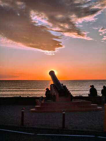 Um canhão está em uma silhueta contra um pôr do sol deslumbrante sobre o oceano, com o sol perfeitamente alinhado em sua boca. As pessoas se sentam pacificamente, apreciando a vista.