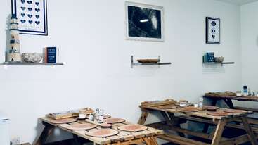 Esta imagen muestra un pequeño y acogedor comedor con mesas de picnic de madera, manteles individuales y condimentos, decorado con arte mural de temática náutica y sencillas estanterías de pared.