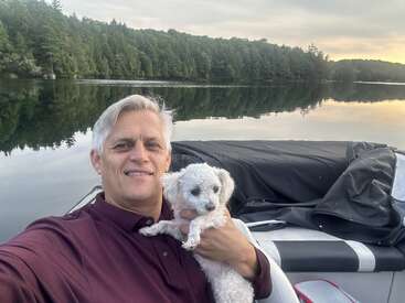 Un homme vêtu d'une chemise marron sourit en tenant un petit chien blanc sur un bateau. Un lac calme et un rivage boisé sont visibles à l'arrière-plan.