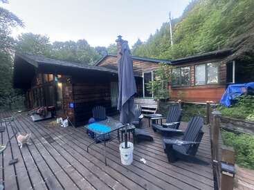 Une cabane en bois confortable se trouve dans une forêt luxuriante. La terrasse spacieuse est équipée de chaises Adirondack, d'une table, d'un parasol fermé et d'un chat qui explore les environs.