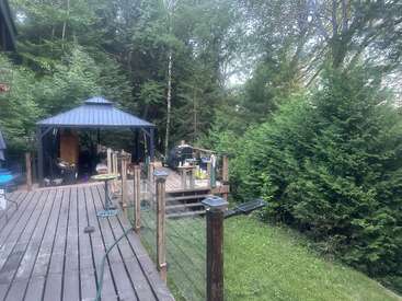 Une terrasse en bois avec un gazebo et un coin grillade est entourée d'arbres et de buissons verdoyants, créant un cadre extérieur paisible et naturel pour se détendre ou se réunir.