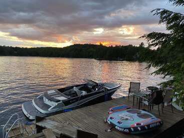Scène sereine au bord d'un lac au coucher du soleil, comprenant un quai avec un bateau, des chambres à air, des meubles de terrasse, des arbres et une eau calme reflétant un ciel spectaculaire.