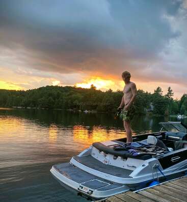 Un homme torse nu se tient à l'arrière d'un bateau, amarré sur un lac calme, alors que le soleil se couche derrière des arbres verts et des nuages spectaculaires.