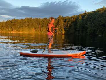 Une femme se tient sur une planche à pagaie, profitant d'une eau calme au coucher du soleil. Elle porte un gilet de sauvetage, entourée d'un rivage boisé et de reflets dorés.
