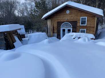 Une cabane en bois est entourée d'une neige épaisse et intacte. Les arbres à l'arrière-plan sont dénudés et tout est recouvert d'un épais manteau blanc.