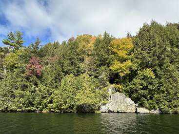 Une colline boisée luxuriante s'élève au-dessus d'un lac calme, avec un feuillage vert, jaune et rouge. De gros rochers bordent l'eau.