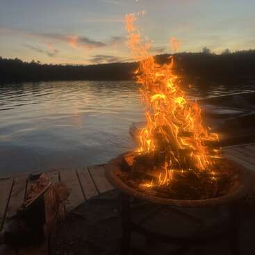 Ein loderndes Lagerfeuer brennt auf einem hölzernen Steg an einem friedlichen See bei Sonnenuntergang, während sich sanfte Wolken und Bäume gegen den Abendhimmel abzeichnen.