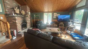 Das gemütliche Wohnzimmer ist mit Ledersofas, einem gemauerten Kamin, einer Holzdecke, großen Fenstern, einem Fernseher, Katzenmöbeln, Kunstwerken und einem rustikalen Couchtisch ausgestattet und versprüht Hüttencharme.