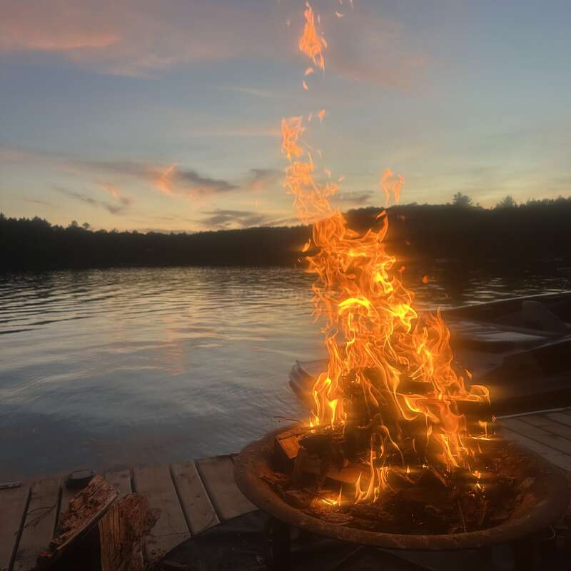 Un feu de camp brûle intensément sur un quai en bois au bord d'un lac paisible au coucher du soleil, avec des nuages doux et des arbres se silhouettant dans le ciel du soir.