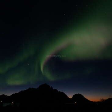 The image depicts the Northern Lights, also known as the Aurora Borealis, in a dark sky above a mountain range. The lights are green and red.