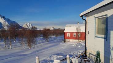 In einer verschneiten Landschaft sieht man ein rotes und ein weißes Haus, kahle Bäume, Berggipfel im Hintergrund und einen klaren blauen Himmel, der in Sonnenlicht getaucht ist.