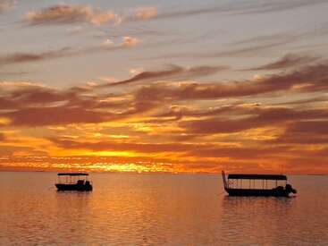 Zwei Boote treiben ruhig auf dem ruhigen Wasser unter einem leuchtenden, orangefarbenen Himmel bei Sonnenuntergang. Zarte Wolken ziehen über den Horizont und spiegeln sich wunderschön in der ruhigen See.