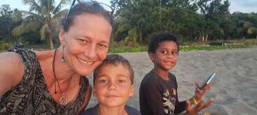 Eine Frau und zwei Jungen sitzen an einem Sandstrand und lächeln in die Kamera. Palmen und üppiges Grün füllen den Hintergrund. Ein Junge hält ein Telefon in der Hand.