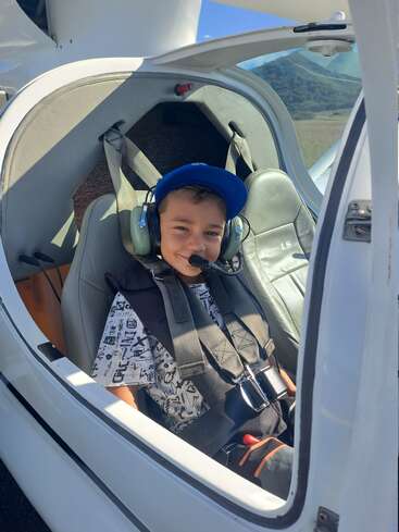 Ein lächelnder Junge sitzt in einem kleinen Flugzeugcockpit, trägt ein Headset, eine blaue Mütze und einen Sicherheitsgurt und ist bereit für ein aufregendes Flugabenteuer.