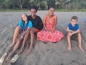 Vier Kinder sitzen zusammen am Sandstrand, zwei Mädchen in Kleidern lächeln, ein Mädchen mit Zöpfen entspannt und ein Junge im blauen Hemd schaut zur Seite.