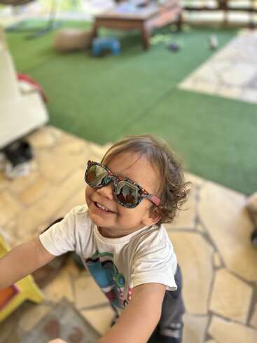 Ein fröhliches Kleinkind, das eine bunte Sonnenbrille trägt, lächelt strahlend und reckt sich nach oben. Er steht auf einer Terrasse mit grünem Rasen und Spielzeug im Hintergrund und strahlt vor Glück.