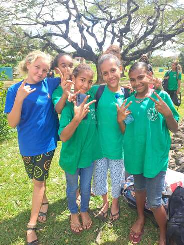 Fünf fröhliche Mädchen in grünen und blauen Hemden posieren im Freien. Sie lächeln und machen Friedenszeichen. Sie genießen einen sonnigen Tag unter den Bäumen in einem Park.
