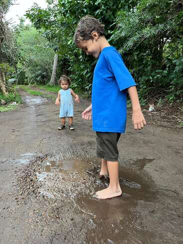 Zwei Kinder spielen draußen auf einem schlammigen Weg. Das ältere Kind planscht vergnügt barfuß in einer Pfütze, während das jüngere in der Nähe steht und neugierig zusieht. Üppiges Grün umgibt sie.