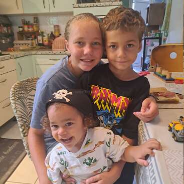 Drei lächelnde Kinder posieren zusammen in einer gemütlichen Küche und umarmen sich voller Freude. Der Jüngste trägt einen Piratenhut, und Spielzeug und Holzklötze schmücken den Tisch daneben.