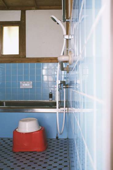 L'image représente une salle de bains avec des carreaux bleus, une douche et un siège de pot rouge, avec une fenêtre et des garnitures en bois, dégageant une atmosphère propre et organisée.