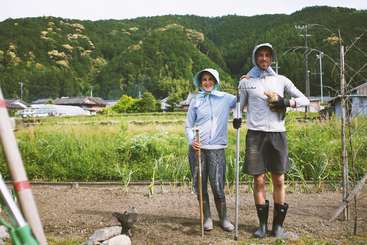 L'image montre un groupe de personnes en train de jardiner, un homme et une femme se tenant ensemble, entourés d'arbres et de buissons, avec un plan d'eau en arrière-plan. La scène est sereine et paisible.