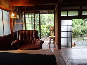 L'image montre une chambre sereine avec un fauteuil en cuir, un lampadaire et un lit, avec une porte coulissante en verre qui mène à un jardin luxuriant avec un poulet qui se promène librement à l'extérieur.