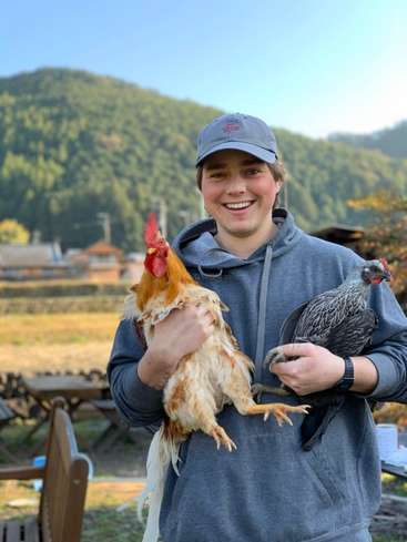 L'image représente un homme tenant deux poulets, l'un blanc et l'autre noir, sur fond de paysage montagneux.