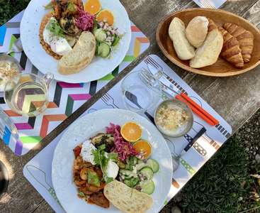 L'image représente une table en bois avec deux assiettes de nourriture, un bol de pain et un verre d'eau, préparés pour un repas par une journée ensoleillée.