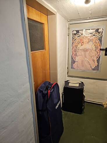 Une petite pièce faiblement éclairée avec une porte en bois, deux grands sacs, des murs blancs, un sol vert et une affiche de style Art nouveau sur le mur.