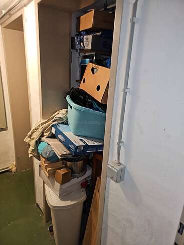 Un petit espace de stockage rempli d'objets divers : des boîtes, un panier à linge, des cartons, des fournitures ménagères et des objets divers empilés de façon chaotique dans un placard étroit.