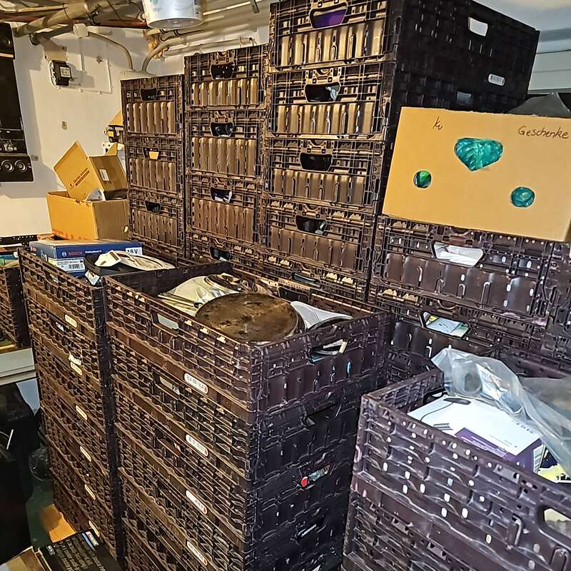 L'image montre un local de stockage encombré de nombreuses caisses en plastique marron empilées, de boîtes en carton et d'objets divers. Des tuyaux et des équipements électriques sont visibles sur les murs.