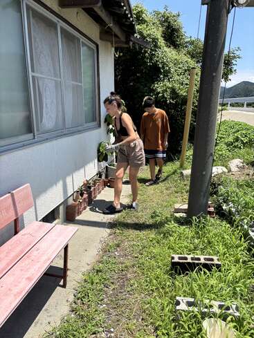 Zwei Menschen stehen an einem sonnigen Tag vor einem Haus. Der eine gießt die Topfpflanzen, die an der Wand stehen, während der andere sie beobachtet. Gras und Ziegelsteine umgeben sie.