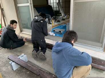 Drei Personen sitzen und knien vor einem Haus und arbeiten gemeinsam an einer Schiebetür. Es sind Werkzeuge und Baumaterialien zu sehen, die auf ein Heimwerkerprojekt hindeuten.