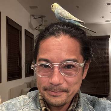 A man with clear-framed glasses and long dark hair smiles indoors. A light blue and yellow parakeet is perched comfortably on top of his head.