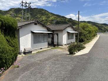 A small white house sits quietly by a roadside, surrounded by lush green hills, power lines, blue sky, and some shrubs and flowers in the foreground.