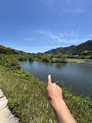 A hand points toward a serene river surrounded by lush greenery and mountains under a clear, bright blue sky with wispy clouds on a sunny day.