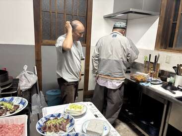 Two men prepare food in a small kitchen with wooden window frames. Several plates of fresh ingredients and sliced meat are arranged on the counter nearby.