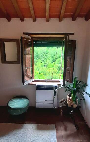 A cozy room with terracotta ceiling beams features an open window overlooking lush greenery, a potted plant, a heater, a green stool, and a decorative rug.