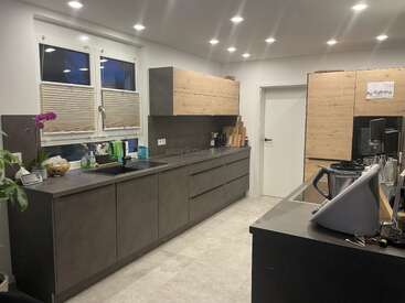 Esta cozinha moderna apresenta armários cinza elegantes e detalhes em madeira, sob uma iluminação embutida brilhante. Os eletrodomésticos, as plantas e os utensílios estão bem organizados, criando um espaço limpo e funcional.